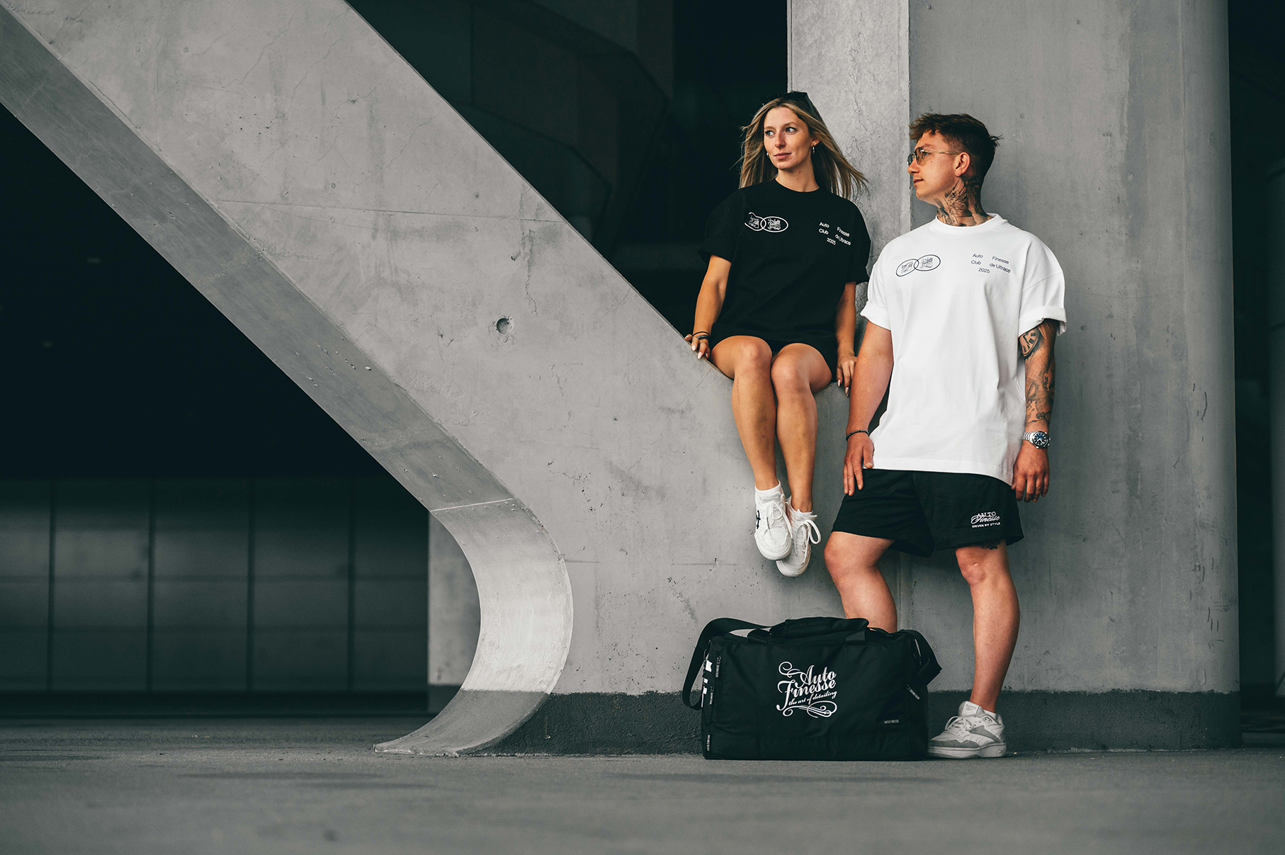 Two people standing next to a black duffel bag with visible branding in an urban setting.