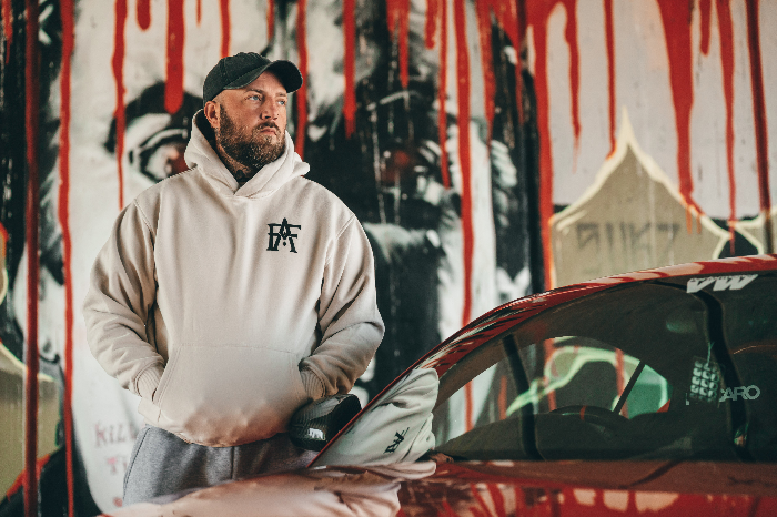 Man wearing a beige hoodie with a logo, standing next to a red car against a graffiti-covered wall.