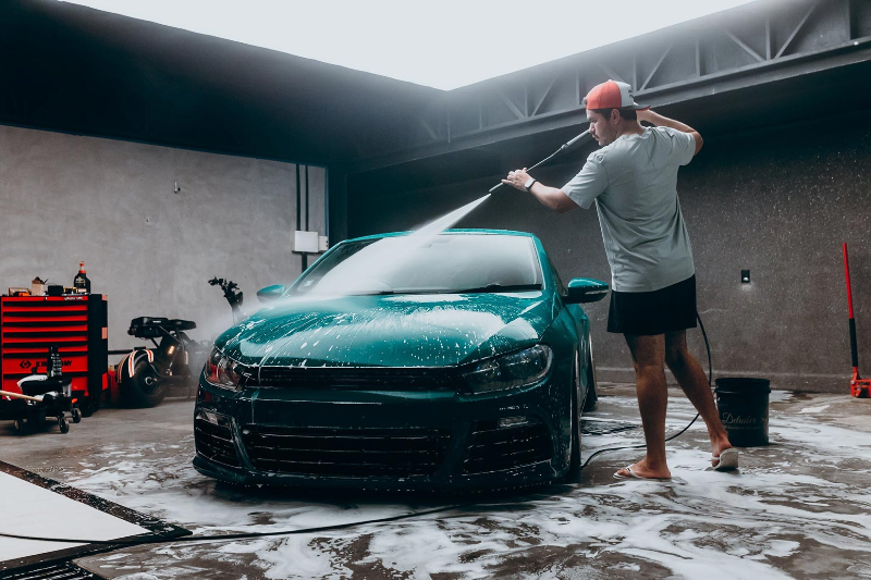 Person washing a green car with a high-pressure water hose in an outdoor setting.
