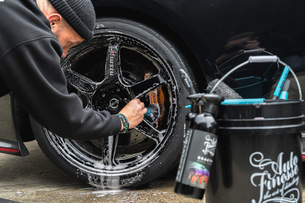 Person washing a car wheel with a brush, using a black bucket and cleaning products.