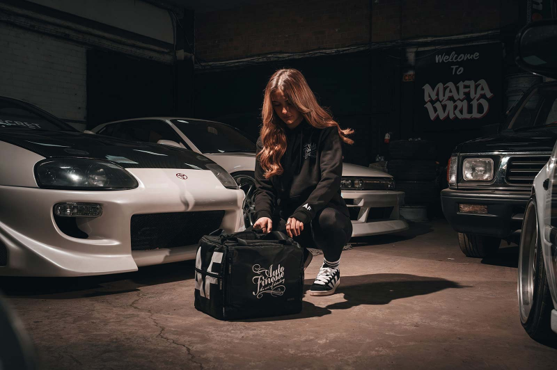 Person with long hair in a garage with cars and a bag featuring 'Auto Finesse' branding.