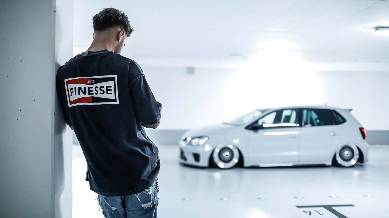Person wearing a black hoodie with 'Finesse' logo looking at a white car in a garage.